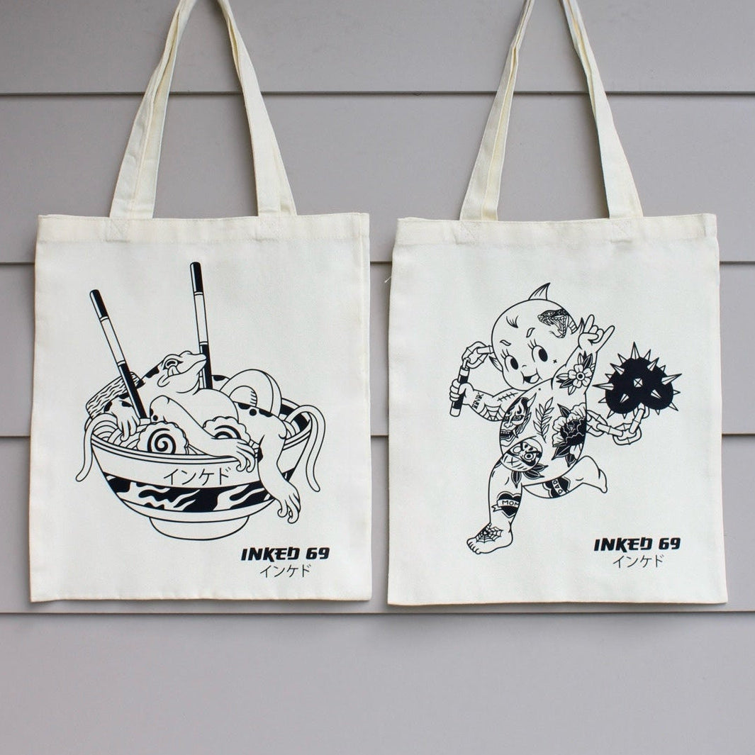 Two off-white canvas tote bags hanging against a light gray horizontal-slat wall. Each bag has a black line-art illustration and the text 'INKED 69' printed near the bottom.