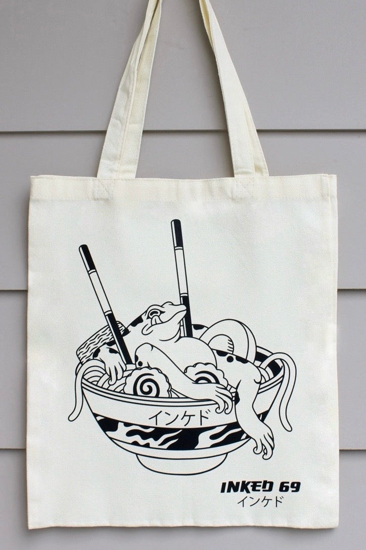 An off-white canvas tote bag with long handles, featuring a black line art illustration of a ramen bowl with a cat and the text 'INKED 69' and Japanese characters, hanging against a gray wood-paneled wall.