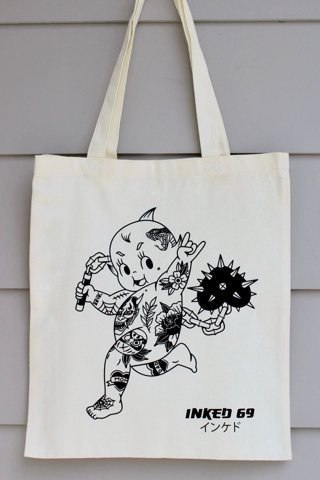 An off-white canvas tote bag with long handles, featuring a black line art illustration of a tattooed character and the text 'INKED 69' printed on the front, hanging against a gray wood-paneled wall.