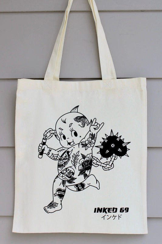 An off-white canvas tote bag with long handles, featuring a black line art illustration of a tattooed character and the text 'INKED 69' printed on the front, hanging against a gray wood-paneled wall.