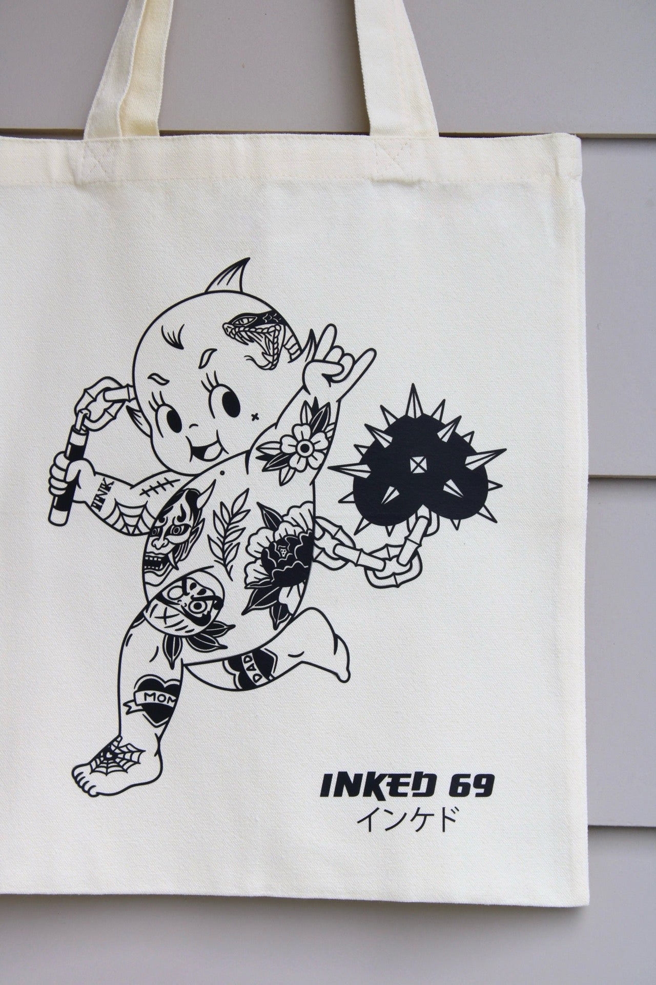An off-white canvas tote bag featuring a black line art illustration of a tattooed character and the text 'INKED 69' with Japanese characters below, shown hanging against a gray paneled wall.