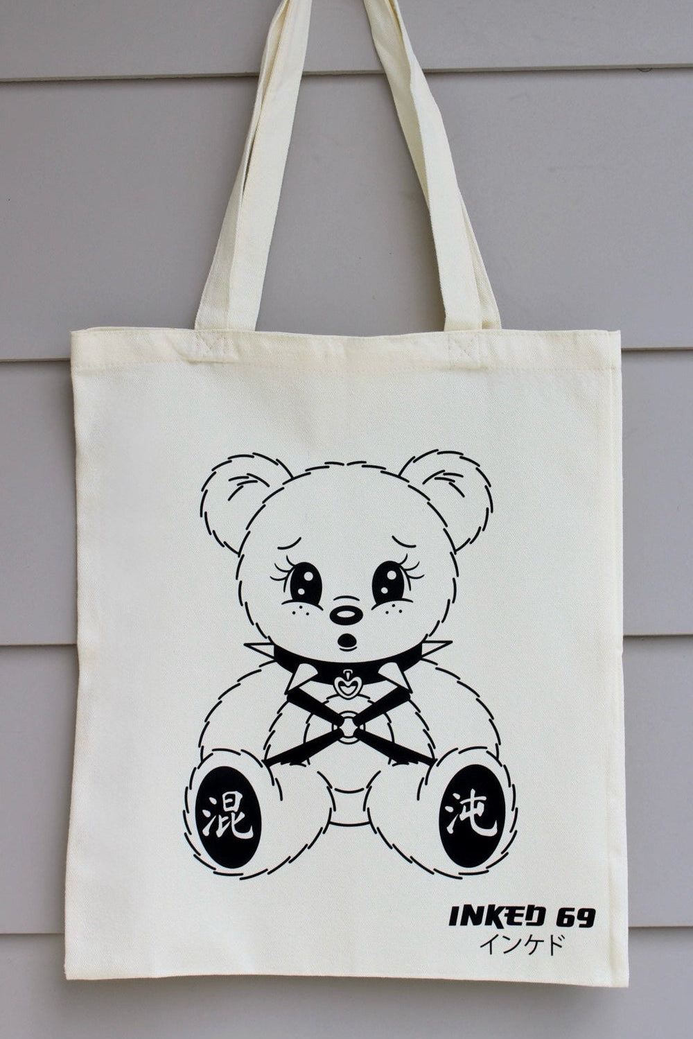 An off-white canvas tote bag hanging against a gray horizontal siding wall. The bag features a black line art illustration of a teddy bear wearing a black and white striped shirt, with Japanese characters on its feet and 'INKED 69' printed below.