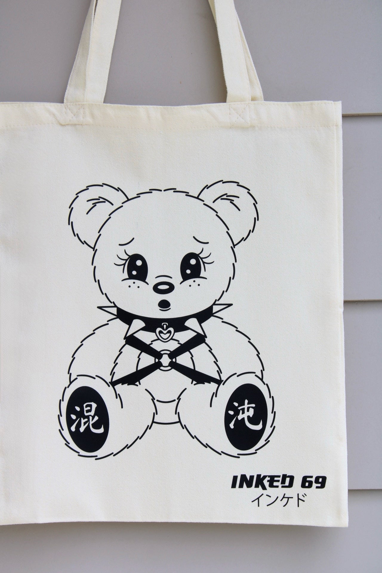 An off-white canvas tote bag featuring a black line art illustration of a teddy bear with Japanese characters on its paws and the text 'INKED 69' and 'こтон' printed below. The bag is hanging against a light gray paneled wall.