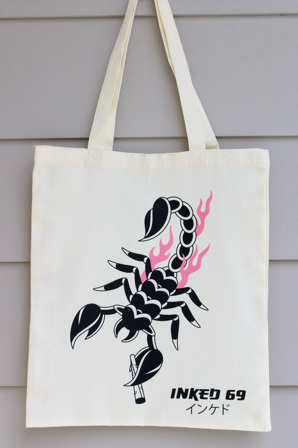An off-white canvas tote bag hanging against a light gray wood-paneled wall. The bag features a black scorpion graphic with pink flames and the text 'INKED 69' printed on the front.