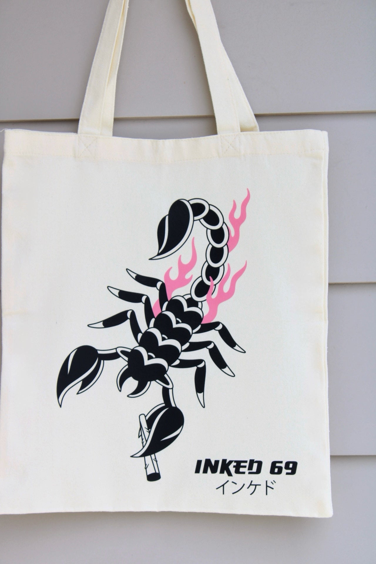 An off-white canvas tote bag hanging against a light gray paneled wall, featuring a black scorpion graphic with pink flames and the text 'INKED 69' printed below.