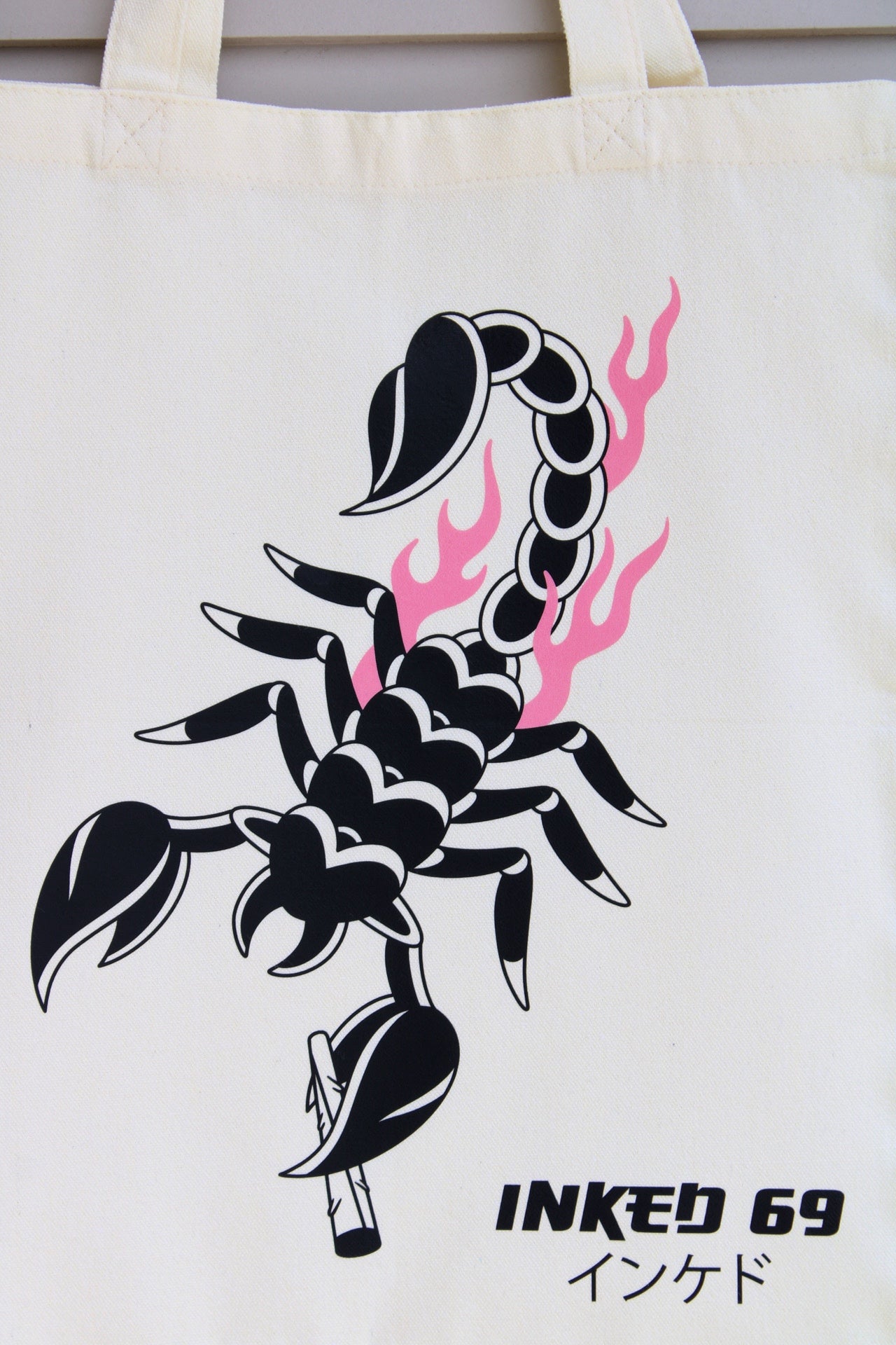 An off-white canvas tote bag featuring a black scorpion graphic with pink flames and the text 'INKED 69' printed on the front.