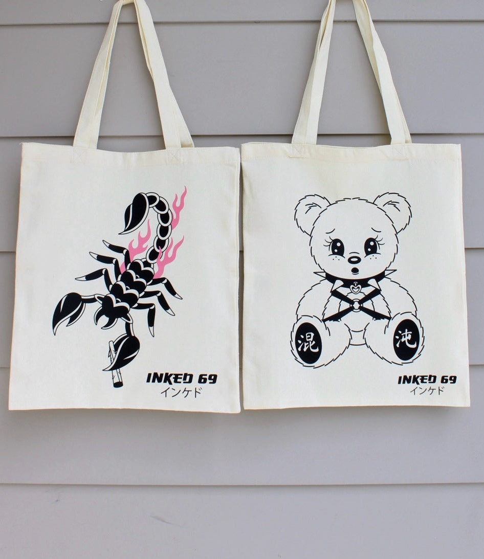 Two off-white canvas tote bags hanging against a light gray wood-paneled wall. The left bag features a black scorpion graphic with pink flame accents and the text 'INKED 69' with Japanese characters below.