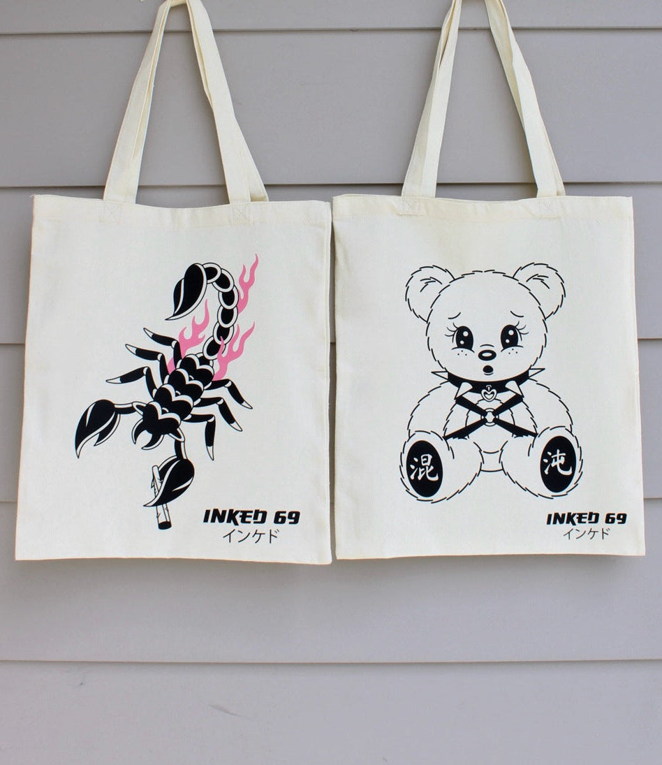 Two off-white canvas tote bags hanging against a light gray wood-paneled wall. The left bag features a black scorpion graphic with pink flame accents and the text 'INKED 69' with Japanese characters below.