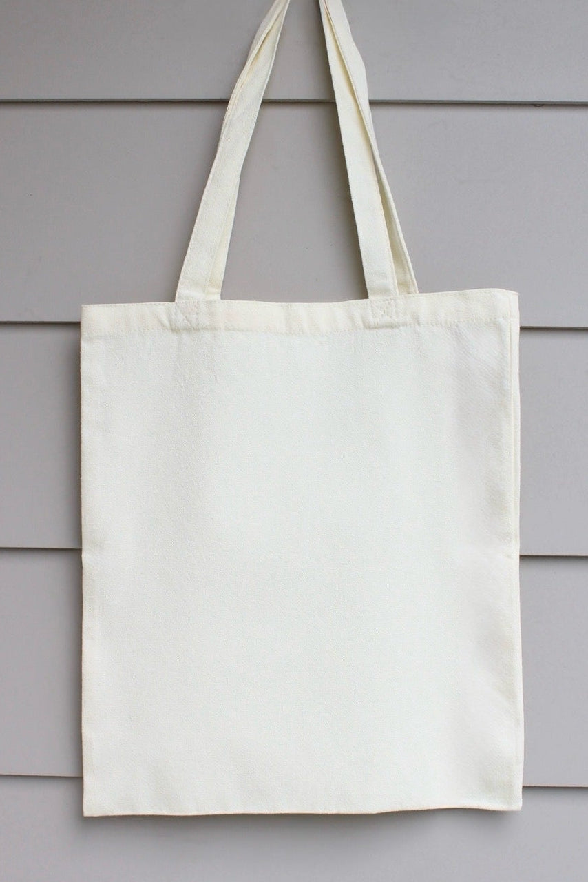 An off-white canvas tote bag with two shoulder straps, hanging against a light gray horizontal siding wall.