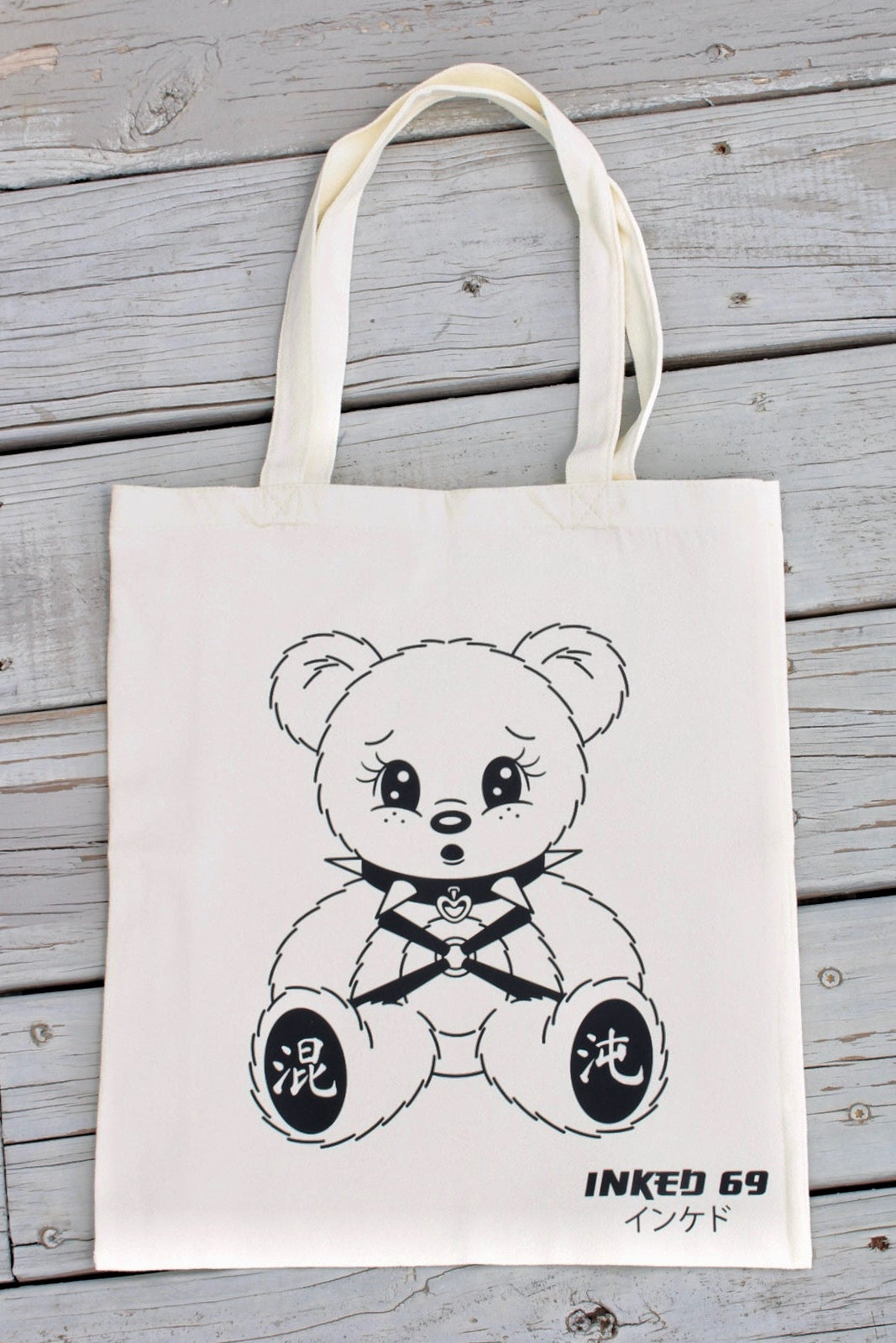 An off-white canvas tote bag with a black line art illustration of a teddy bear wearing a bow tie, displayed flat on a gray wooden plank surface. The bag features Japanese characters on the bear's paws and 'INKED 69' with Japanese text near the bottom right.