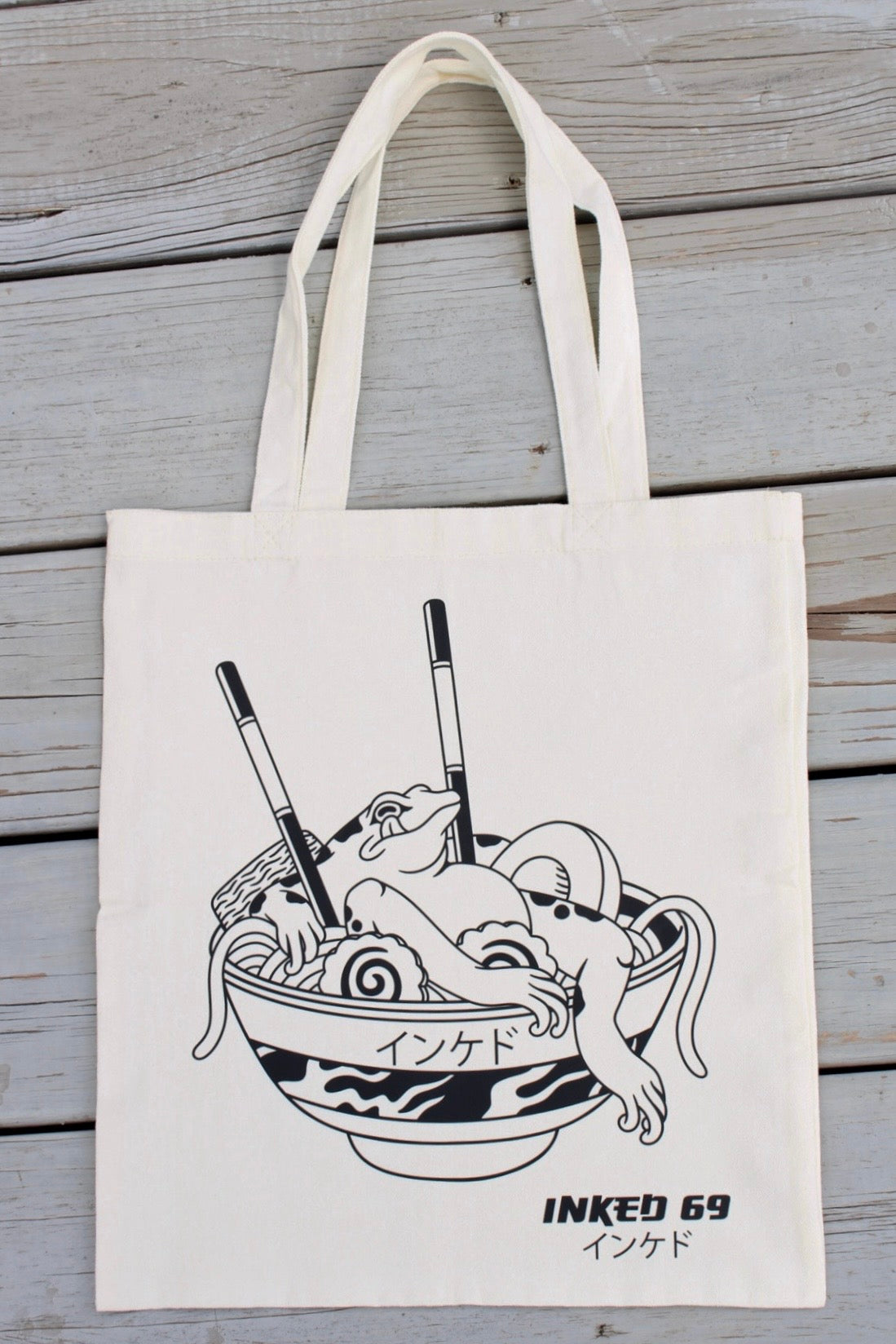 An off-white canvas tote bag with black line art of a ramen bowl and the text 'INKED 69' and 'こいじおす' on a weathered gray wooden plank background.