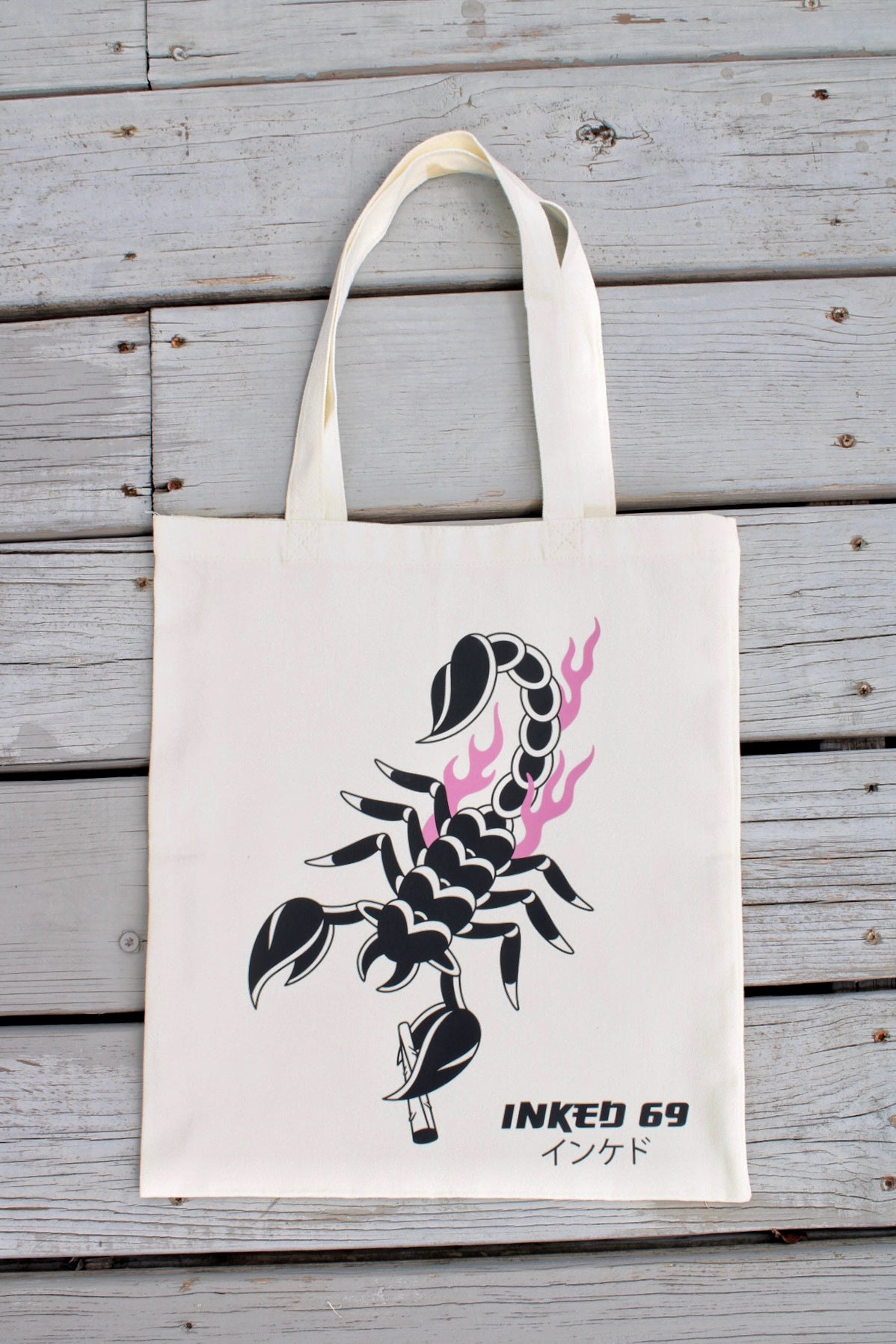 An off-white canvas tote bag with a black scorpion graphic and pink flame accents, featuring the text 'INKED 69' and Japanese characters, displayed against a gray wooden plank background.