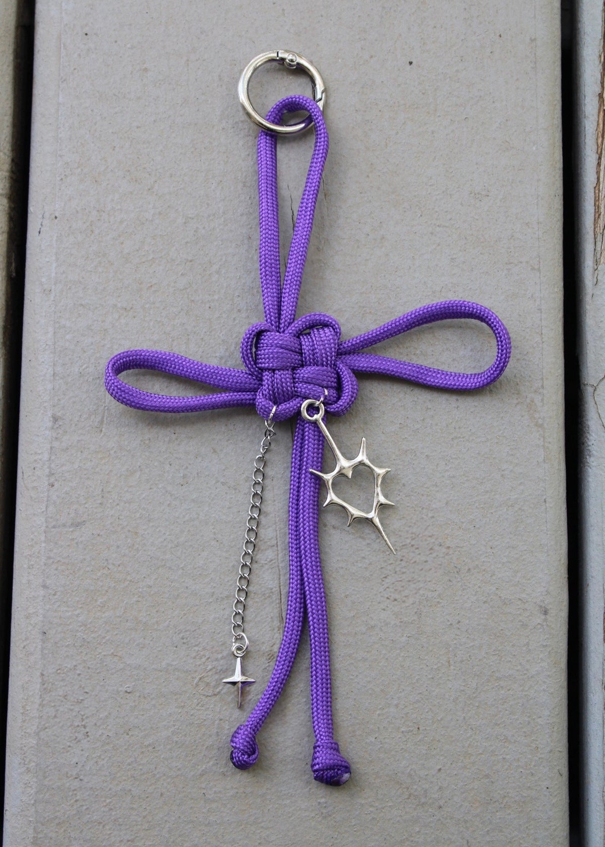 A purple paracord keychain charm shaped like a stylized cross with a silver metal key ring, chain, and starburst and lightning bolt charms, displayed on a gray wooden background.