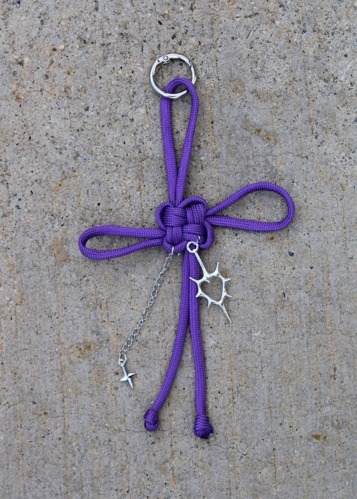 A purple paracord keychain or bag charm with a silver metal ring, keyring, and sunburst charm, laid flat on a textured gray concrete surface.