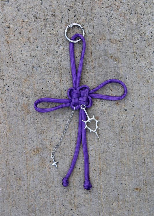 A purple paracord keychain or bag charm with a silver metal ring, keyring, and sunburst charm, laid flat on a textured gray concrete surface.