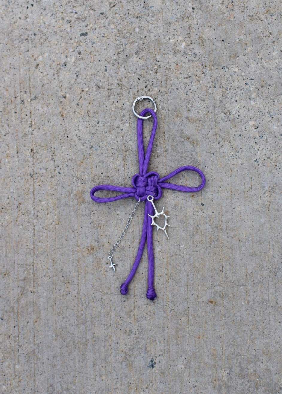 A purple paracord keychain charm with a knotted bow, silver metal ring, and small dangling star and crescent moon charms, placed on a textured concrete surface.