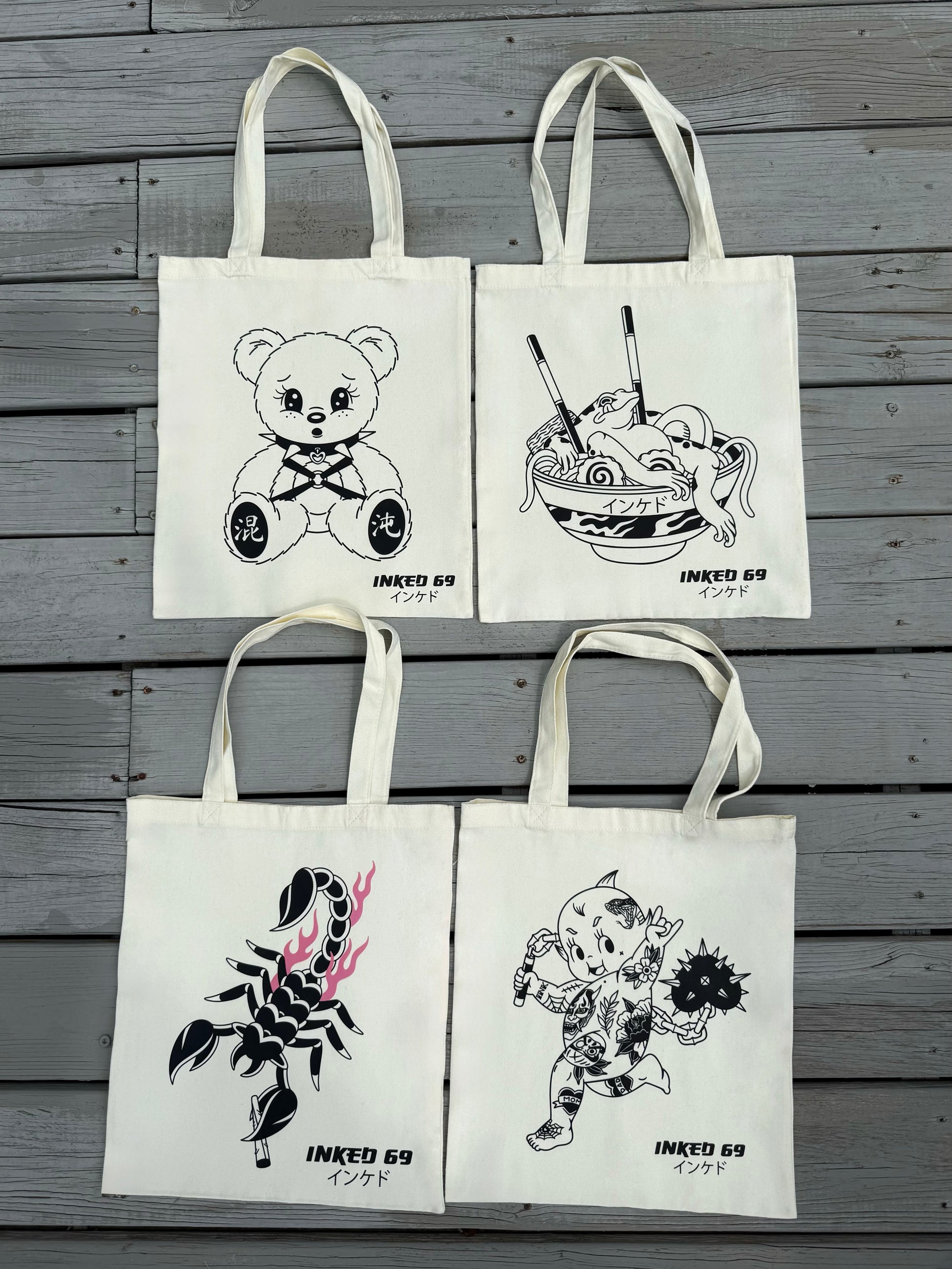 Four off-white canvas tote bags arranged in a 2x2 grid on a gray wooden plank background. Each bag features a different black line-art illustration: a teddy bear, ramen, a scorpion with pink flames, and a cartoon character, all labeled 'INKED 69'.